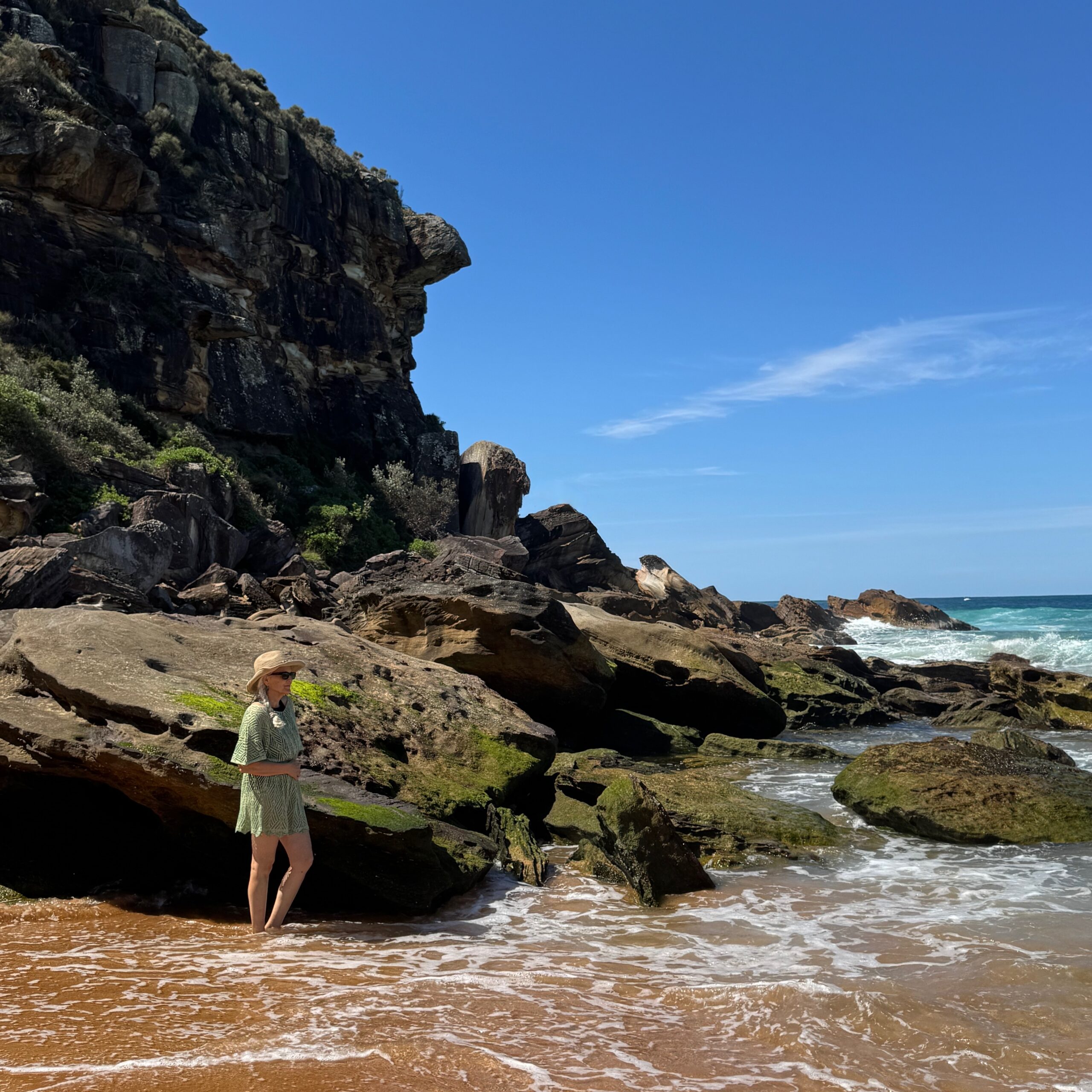 The author stands with her feet in the surf, rocks behind her, in Palm Beach, Australia